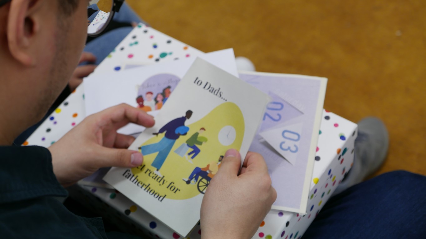 A man holds a book with an illustration of 3 men holding babies. It reads 'to Dads, get ready for fatherhood'